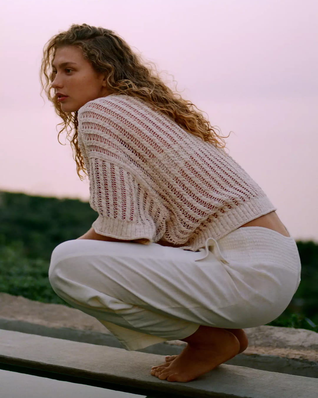 Woman crouching outdoors at sunset, wearing cream open-knit sweater and white trousers, lifestyle shot with pink sky background