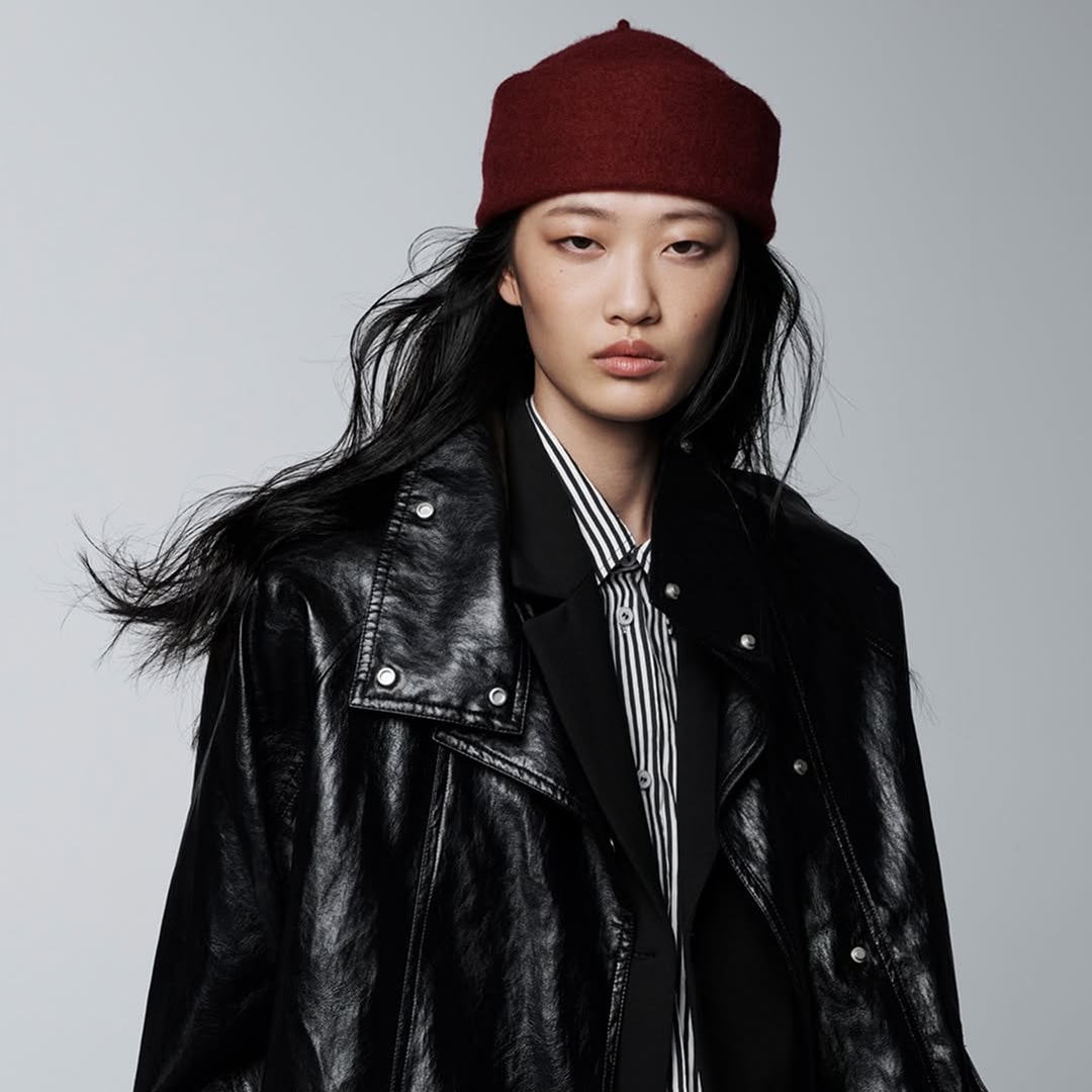 Model wearing a red beanie, black leather jacket, and striped shirt against a gray background